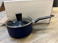 Blue T-Fal saucepan with black handle and matching glass lid, placed on a white box, showing side view