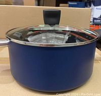 Blue T-FAL Dutch oven with glass lid, showing side view and black Bakelite side handle, placed on cardboard surface.
