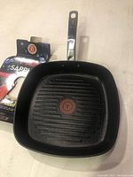 Square black non-stick grill pan with ridged cooking surface, stainless steel handle showing T-Fal logo, and red Thermo-Spot heat indicator in center. Original product packaging partially visible nearby.