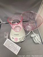 Conair facial sauna unit with pink warm steam bowl, clear cover, power cord, and instruction manual on display against a gray backdrop.