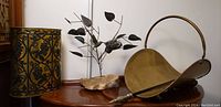 Photo showing the brass firewood holder with handle, the antique brass bucket and the decorative stone base with metal bird sculpture on a wooden surface.