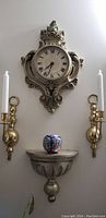 Full view of antique wall clock flanked by two bronze candle sconces with small decorated vase on wall shelf below