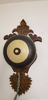 Front view showing black and beige circular metal bell mounted on decorative wooden plaque with floral and leaf carving details.