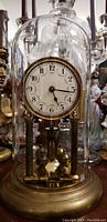 Full view of the glass dome clock showing the porcelain clock face and brass base, housed within a clear glass dome.