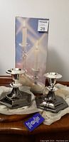 Both silver candle holders positioned in front of original product box packaging, showing new condition with tissue paper, one candle included in photo.