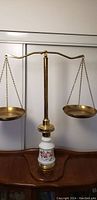 Front view of the antique balance scales showing the brass beam and pans atop a floral porcelain base on a wooden surface.