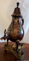 Side angle view of antique hand painted samovar showing scenic painting, curved brass legs, and black dome lid.