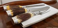 Close-up of three-piece silver serving set with carved antler handles displayed on white porcelain tray with silver rim.