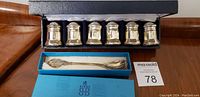 Set of six silver shakers in fitted black box on wooden surface, with blue box containing silver spoon beside it.