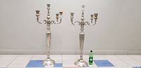 Two large silver plated candelabras side by side with a soda bottle for scale.