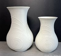 Two white ceramic Kiser vases with raised dot patterns creating circular and flowing wave designs, shown side by side on a grey surface against dark background.
