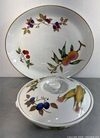 Covered casserole dish in front of matching oval serving platter, both with fruit botanical patterns and gold trim.
