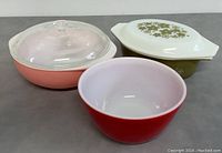 Three vintage Pyrex bowls of different sizes arranged on a table surface, pink bowl with lid, red bowl without lid, olive green bowl with floral lid.