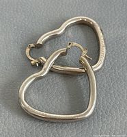 Pair of overlapping heart-shaped silver hoop earrings on gray textured surface.