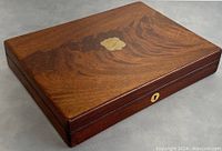 Closed wooden box with rich wood grain, decorative gold-tone emblem on lid and brass escutcheon keyhole on front, shown from a side angle.