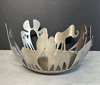 Brushed stainless steel bowl with artistic cutouts shaped like animals placed on gray surface with black background shown at angle.