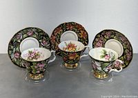 Front view of three Royal Albert teacups with matching floral saucers arranged side by side. Each cup has gold trim and a unique flower pattern on a black background.