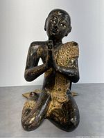 Front view of kneeling monk statue with hands clasped in prayer and detailed gilt patterns on robe.