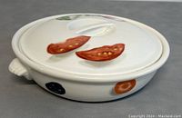 Front angle showing the white porcelain casserole dish with painted tomato slices on the lid and various vegetables around the body, including blue fruit/vegetable and orange rings.
