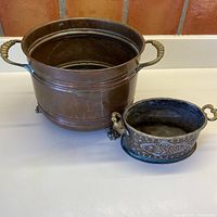 Wide shot showing both copper pots side by side on a white surface with a brick background, highlighting size difference and design elements.