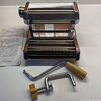 Front and side view of the Beaumark pasta maker with crank handle, clamp, and instruction booklet laid on table.