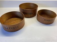 Set of three round teak wood bowls labeled Pretzels, Potato Chips, and Peanuts placed next to each other.