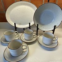 Photo showing the oval platter in the back with a large serving bowl to the right, cups and matching saucers arranged in front. The set has a cream base with cinnamon speckles and cinnamon textured edge detail.