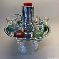 Front view showing chrome rotating plate holding six shot glasses with colored bands and central chrome pump dispenser with red top button.