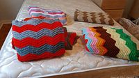 Four handmade afghan blankets folded and laid on a mattress, showing different chevron zigzag crochet color patterns.