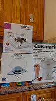 Three boxed appliances on a kitchen counter: Salton portable cooktop, Salter kitchen scale, Cuisinart ice cream maker