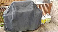 Weber grill covered by vinyl cover on backyard deck with propane tanks