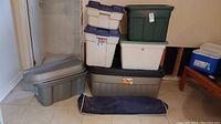 Overview photo showing stack of plastic storage bins of various colors and sizes including Rubbermaid bins, and navy blue vinyl storage bag on floor.