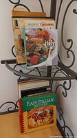 Five assorted cookbooks stacked on a metal shelf. Front book titled 'Easy Italian Recipes' with a photo of a pizza on the cover. Other visible titles include 'A Little Taste of Italy' and '100 Best Good Housekeeping Italian'.