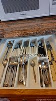 Drawer filled with mixed flatware including forks, spoons, and knives with white and black handles arranged in organizer slots.