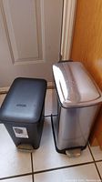 Photo showing two garbage cans side by side; one black plastic and one stainless steel, both with foot pedals and lids.