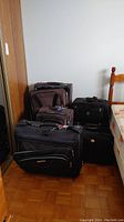 Five pieces of luggage including computer bag, suit bag, and suitcases in black and dark colors grouped near a wall and a chair.