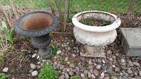 Two planters side by side outside on rocky ground near a chain link fence: one is a dark brown cast iron urn planter and the other is a large white ceramic planter with cracked paint on a pedestal base.