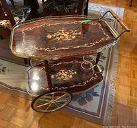 Front angle view of two-tier cart showing woodgrain laminate shelves, floral motif, brass gallery rail and spoked wheels.