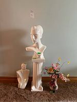 Three white ceramic busts, white pedestal, and vase with faux flowers arranged against wall and carpeted floor.