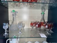 Two decanter sets with matching glasses displayed on shelves. Left is clear crystal decanter and glasses, right is red-tinted set.