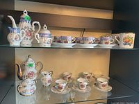 Two shelves displaying two demitasse sets and three mugs, featuring porcelain cups, saucers, coffee pots, creamers, and sugar bowls with detailed decorative scenes and gold accents.