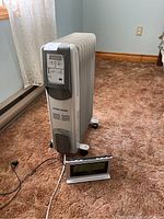Black & Decker radiator heater positioned on a carpeted floor next to a curtain and wall, with power cord visible, and digital clock resting on the floor in front of the heater.
