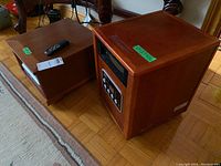 Two portable space heaters with wood-like finish and plastic casters on floor next to wooden furniture.