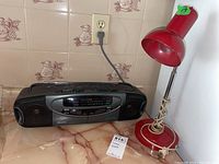 Black Memorex Mp2127 cassette player radio with various buttons and dual speakers, placed on countertop next to red metal desk lamp with cord wrapped around base.