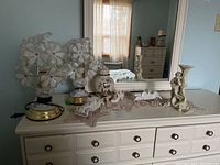 View of two vintage flower lamps, ceramic figure, candle holder, and a lidded jar arranged on a white dresser with lace doily.