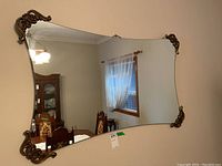 Full view of the large wall mirror mounted on a beige wall reflecting a dining room with wooden furniture and window with lace curtains.
