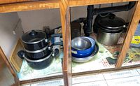 Three black Henri Phillipe pots with lids stacked in cabinet with mixing bowls nearby