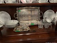 Wide shot of bottom shelf with silver plated trays, ornate serving pieces, and white porcelain plates and bowls with floral patterns