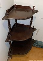 Three-tier wood corner shelf placed against a white corner wall. Showing side and front view with turned posts and finials on top.