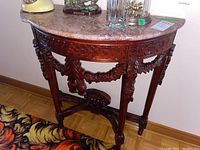 Front and side views of a demi lune table with reddish stone top and intricately carved wood base featuring floral motifs and garlands.
